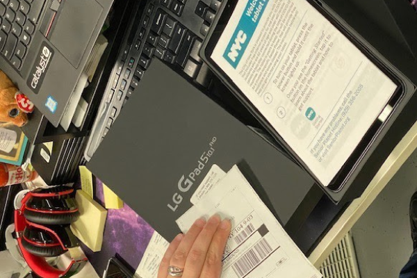 A product box marked with the LG logo and model name of the tablet being distributed, open on a desk, with a folded piece of paper branded with the NYC logo placed inside and a shipping label bring prepared.